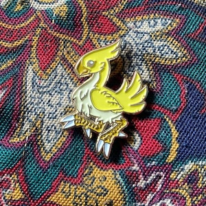 May include: A yellow and white enamel pin of a Chocobo, a bird-like creature from the Final Fantasy video game series. The pin has a gold finish and features a white outline.