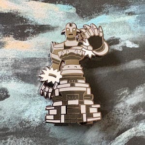 Può includere: Uno spillo in smalto argento e bianco di un golem di pietra con una mano alzata. Il golem è in piedi su una base di blocchi di pietra impilati.