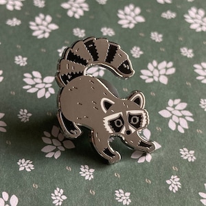 May include: A silver enamel pin featuring a cartoon raccoon with a black and white striped tail. The raccoon is facing forward with its head tilted slightly to the side. The pin has a black outline and white eyes.