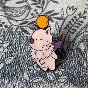 May include: A pink enamel pin featuring a cartoon character with closed eyes, a purple nose, and a fluffy collar. The character has purple bat wings and a yellow orb above its head. The pin has a black outline.