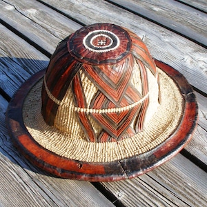 May include: A woven hat with a brown leather brim and a patterned top. The hat is made of natural materials and has a unique design.