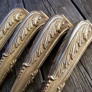 Op de afbeelding: Een set van vier goudkleurige metalen meubelpoten met een sierlijk, verhoogd bladpatroon aan de bovenzijde en een textuur met stippen op het onderste, taps toelopende gedeelte, gekenmerkt door gedetailleerde afwerking en een metaalglans.