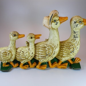 Puede incluir: Un tope de puerta de hierro fundido con cuatro patos en fila. Los patos están pintados de blanco con picos y patas amarillas. El pato más grande tiene un sombrero rosa y negro. El tope de puerta está sobre un fondo blanco.