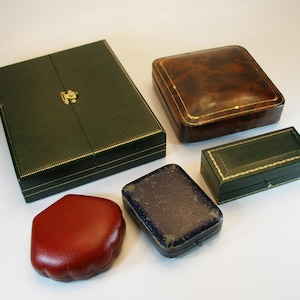 Op de afbeelding: Een assortiment vintage leren sieradendoosjes in verschillende vormen en maten. De doosjes zijn in tinten groen, bruin, rood en blauw, met gouden accenten. De doosjes zijn gesloten en leeg.