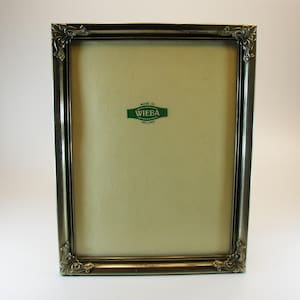 Op de afbeelding: Een rechthoekige fotolijst met een donkere metallic afwerking en sierlijke hoekdetails. De lijst heeft een lichtgekleurde achterkant en een groen label met de tekst "MADE IN WIEBA HOLLAND".