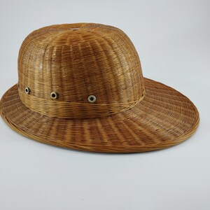 May include: A woven brown hat with a wide brim and a rounded crown. The hat has three small metal eyelets on the front. The hat is made of a light brown material and appears to be made of woven straw or wicker.