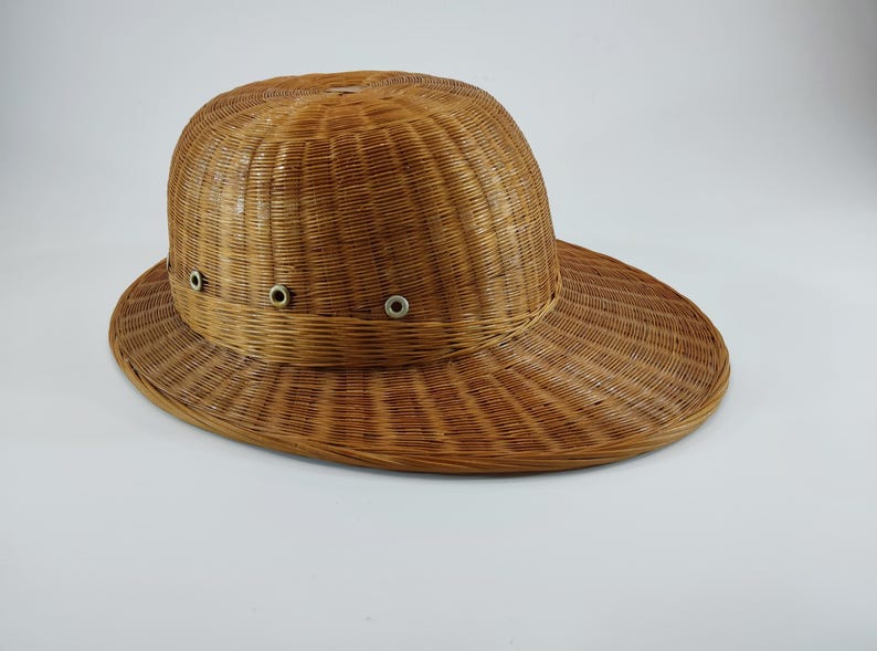 May include: A woven brown hat with a wide brim and a rounded crown. The hat has three small metal eyelets on the front. The hat is made of a light brown material and appears to be made of woven straw or wicker.