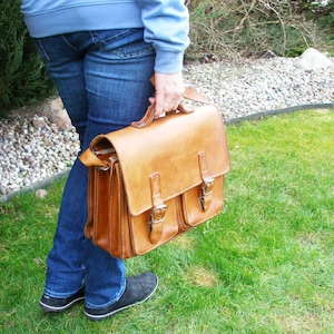 Puede incluir: Un maletín de cuero marrón con dos bolsillos delanteros y un cierre de hebilla. El maletín tiene un asa superior y una correa para el hombro.