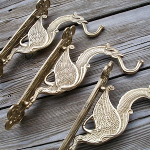 Puede incluir: Tres ganchos de pared de metal dorado con forma de dragones estilizados con alas y detalles ornamentados. Los ganchos están montados en una superficie de madera.