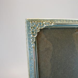 May include: A silver picture frame with an ornate design around the edge. The frame is empty and has a light blue background.