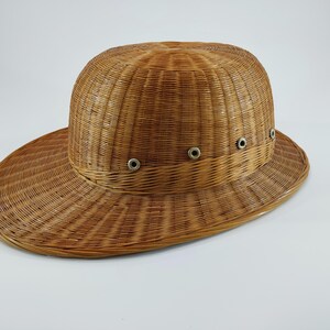 May include: A woven brown hat with a wide brim and a rounded crown. The hat features a series of small, silver-colored eyelets around the crown. The hat is made of a natural, light brown material, likely wicker or straw.