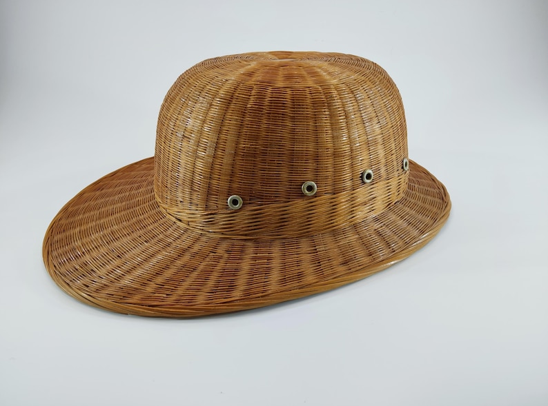 May include: A woven brown hat with a wide brim and a rounded crown. The hat features a series of small, silver-colored eyelets around the crown. The hat is made of a natural, light brown material, likely wicker or straw.