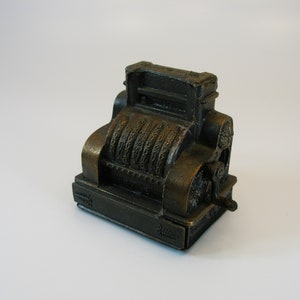 Peut inclure: Un taille-crayon en métal de style vintage en forme de caisse enregistreuse. Le taille-crayon est brun et possède une manivelle sur le côté. Il porte une étiquette "Junior" sur le fond.