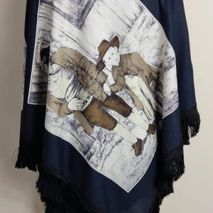 May include: A dark navy blue poncho featuring a large central rectangular graphic of two figures in hats and traditional clothing. The graphic includes the text "Rainer Herzer" and is surrounded by a solid navy border with black fringe trim.
