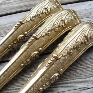 May include: Three gold-toned metal furniture legs with ornate, detailed designs. The legs have a textured surface and a curved shape.