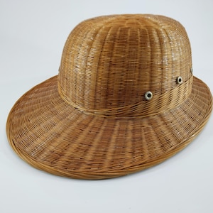 May include: A woven straw hat with a wide brim and a rounded crown. The hat is light brown and features small metal eyelets around the crown. The hat's design is reminiscent of a safari or sun hat.