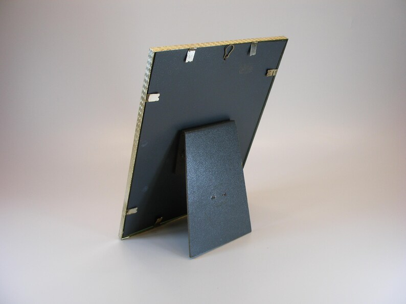 May include: A gold-colored picture frame with a black mat and a stand on the back. The frame is made of a textured material.