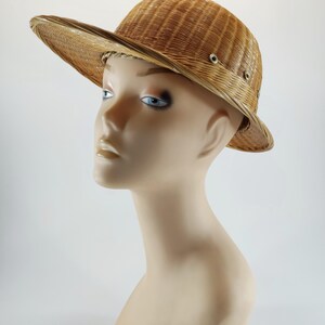 May include: A woven straw hat with a wide brim and a slightly curved crown. The hat is a light tan color and features small metal eyelets around the crown. The hat is displayed on a mannequin head.