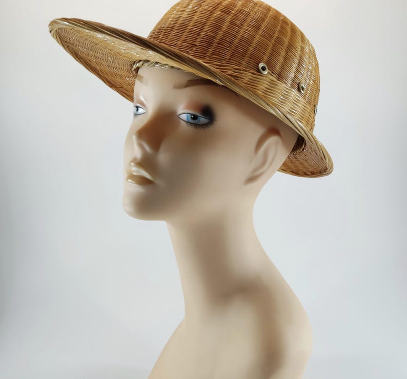May include: A woven straw hat with a wide brim and a slightly curved crown. The hat is a light tan color and features small metal eyelets around the crown. The hat is displayed on a mannequin head.