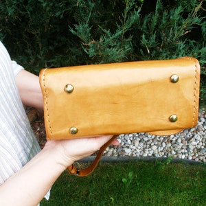 Puede incluir: Un bolso de mano de cuero marr&oacute;n con asa superior y bandolera. El bolso tiene cuatro remaches de lat&oacute;n en la parte inferior.