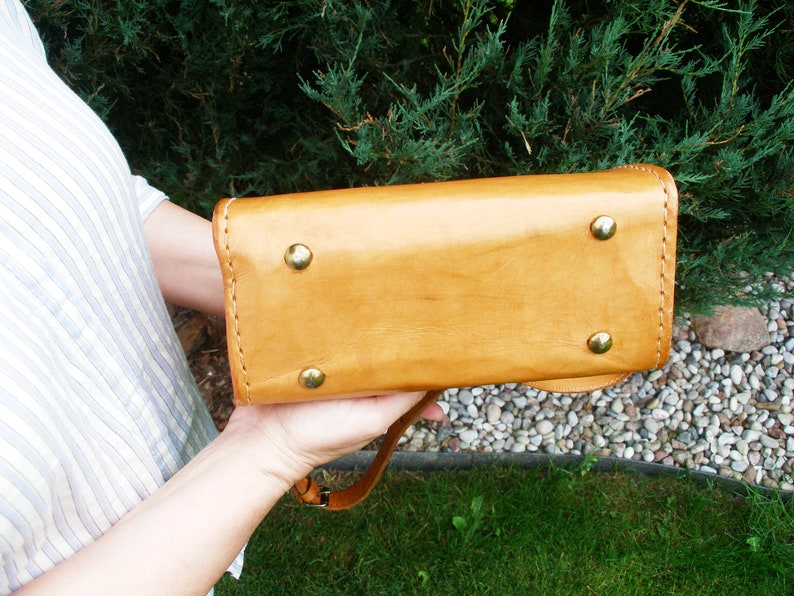 Puede incluir: Un bolso de mano de cuero marr&oacute;n con asa superior y bandolera. El bolso tiene cuatro remaches de lat&oacute;n en la parte inferior.