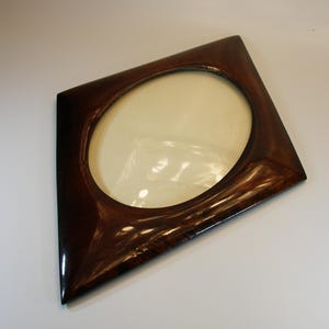 May include: A dark brown, square picture frame with an oval opening. The frame has a glossy finish and a unique, angled design. The frame is empty, with a cream-colored backing visible through the opening. A decorative item for displaying photos or art.