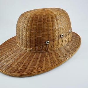 May include: A woven wicker hat with a wide brim and a rounded crown. The hat is a light brown color and features small metal eyelets around the crown. The hat's design suggests it is suitable for sun protection.