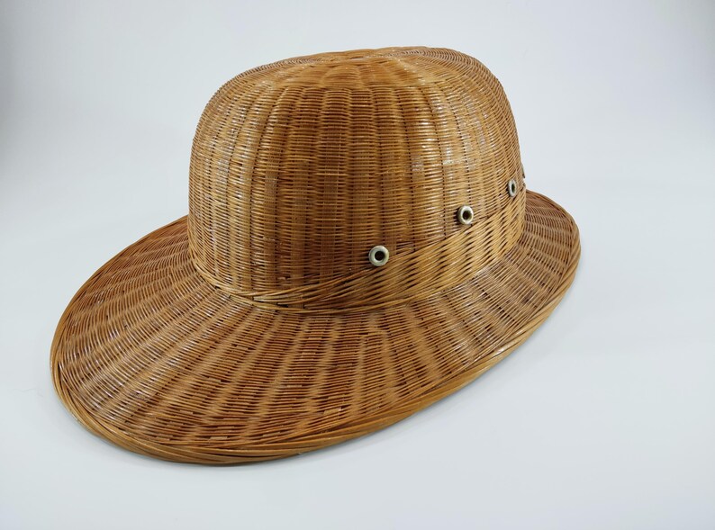 May include: A woven wicker hat with a wide brim and a rounded crown. The hat is a light brown color and features small metal eyelets around the crown. The hat's design suggests it is suitable for sun protection.