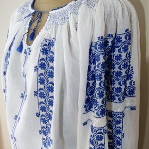 May include: White blouse with blue floral embroidery. The long-sleeved top features detailed blue stitching around the neckline, sleeves, and bodice. A small tassel detail hangs from the neckline. The blouse is made of a lightweight fabric.