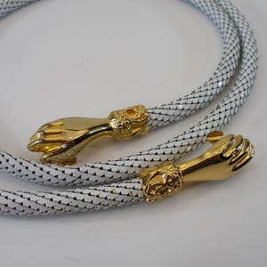 Könnte beinhalten: Ein weißer, geflochtener Kordelgürtel mit zwei polierten, goldfarbenen Metallenden in Form von Händen. Die Hände sind mit Manschetten versehen, die ein Löwenkopfmotiv aufweisen. Die Kordel hat eine schuppenartige Textur und ist kreisförmig gewickelt.