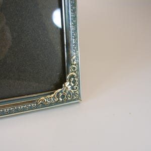 May include: A silver picture frame with an ornate design. The frame has a dark gray mat and a glass front.