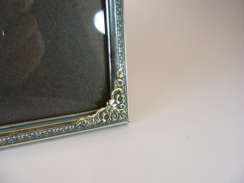 May include: A silver picture frame with an ornate design. The frame has a dark gray mat and a glass front.