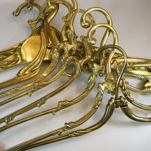 Può includere: Una collezione di grucce in metallo color oro lucido caratterizzate da intricati motivi a volute e ganci curvi. Ogni gruccia presenta una superficie brillante e riflettente con dettagliati motivi in rilievo lungo la struttura.