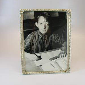 Könnte beinhalten: Ein Schwarzweißfoto eines jungen Menschen, der an einem Schreibtisch sitzt und in ein Notizbuch schreibt. Das Foto ist in einem silbernen Rahmen mit einem verzierten Design gerahmt.