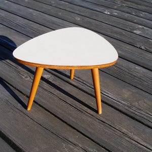 Op de afbeelding: Een kleine bijzettafel met drie poten en een wit, afgerond driehoekig blad met een glad oppervlak. De tafel wordt ondersteund door drie taps toelopende, lichtbruine houten poten die vanuit het midden naar buiten toe uitlopen.