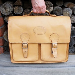 May include: A tan leather briefcase with two front pockets and a brass buckle closure on each pocket. The briefcase has a top handle and a brass clasp.