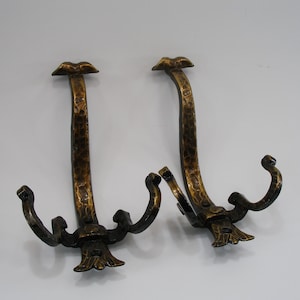 Könnte beinhalten: Zwei antike Wandhaken aus Bronze. Jeder Haken hat einen strukturierten, gebogenen Schaft mit dekorativer Oberseite und zwei gebogenen Haken unten. Die Haken sind dunkelbronze, mit einer leicht gehämmerten Textur.