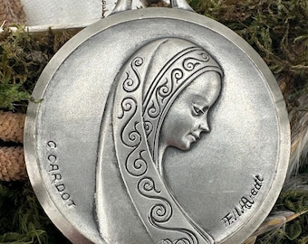 Puede incluir: Un colgante de plata con un relieve detallado de la cara de una mujer con un velo estampado. El colgante está firmado "G. Cardot" y "F.I.L.A. Made".