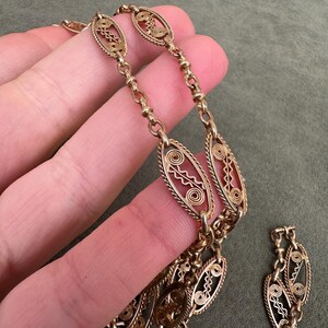 May include: A gold chain necklace with intricate oval links. The links are made of delicate wire that is woven into a pattern of circles and swirls.