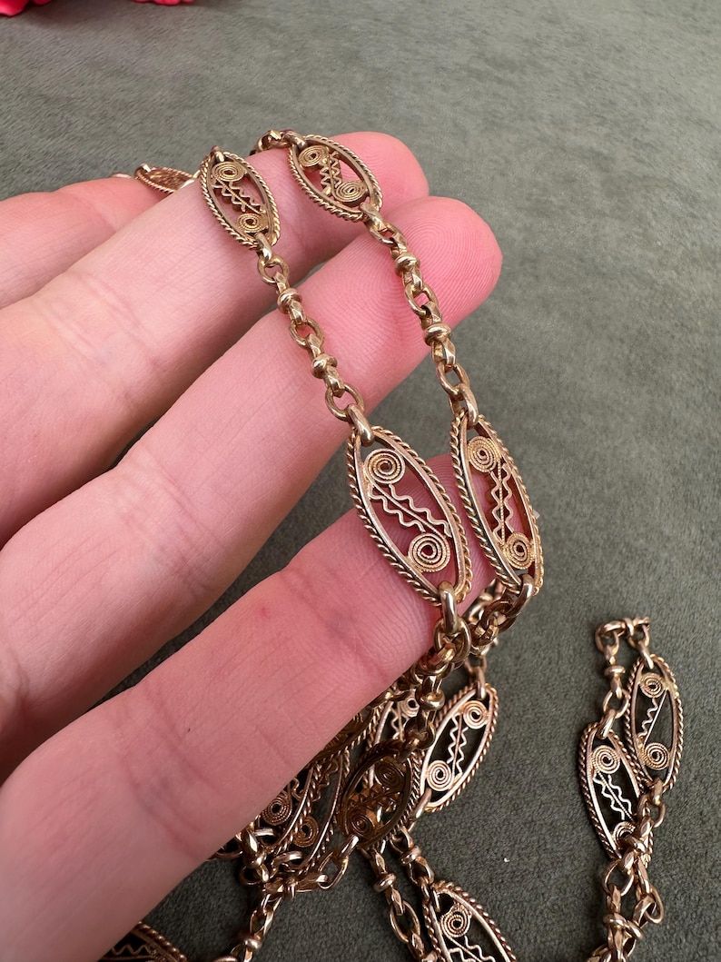 May include: A gold chain necklace with intricate oval links. The links are made of delicate wire that is woven into a pattern of circles and swirls.
