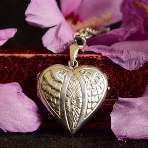 Puede incluir: Un colgante de relicario en forma de corazón de plata con un motivo central de hoja grabada verticalmente, flanqueado por patrones texturizados similares a escamas en ambos lados. El relicario metálico cuelga de una cadena de plata y es brillante.