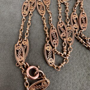 May include: A gold chain necklace with a delicate, intricate design. The chain is made of small, oval-shaped links that are connected by a series of smaller, round links. The necklace has a clasp that is made of a single, larger oval link.