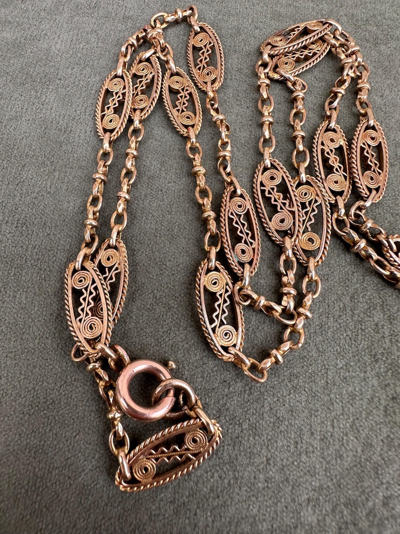 May include: A gold chain necklace with a delicate, intricate design. The chain is made of small, oval-shaped links that are connected by a series of smaller, round links. The necklace has a clasp that is made of a single, larger oval link.