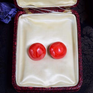 May include: A pair of round, red earrings with a marbled effect, presented in a vintage red jewelry box with a cream-colored satin lining. The earrings are the focal point, showcasing a vibrant color and unique design.