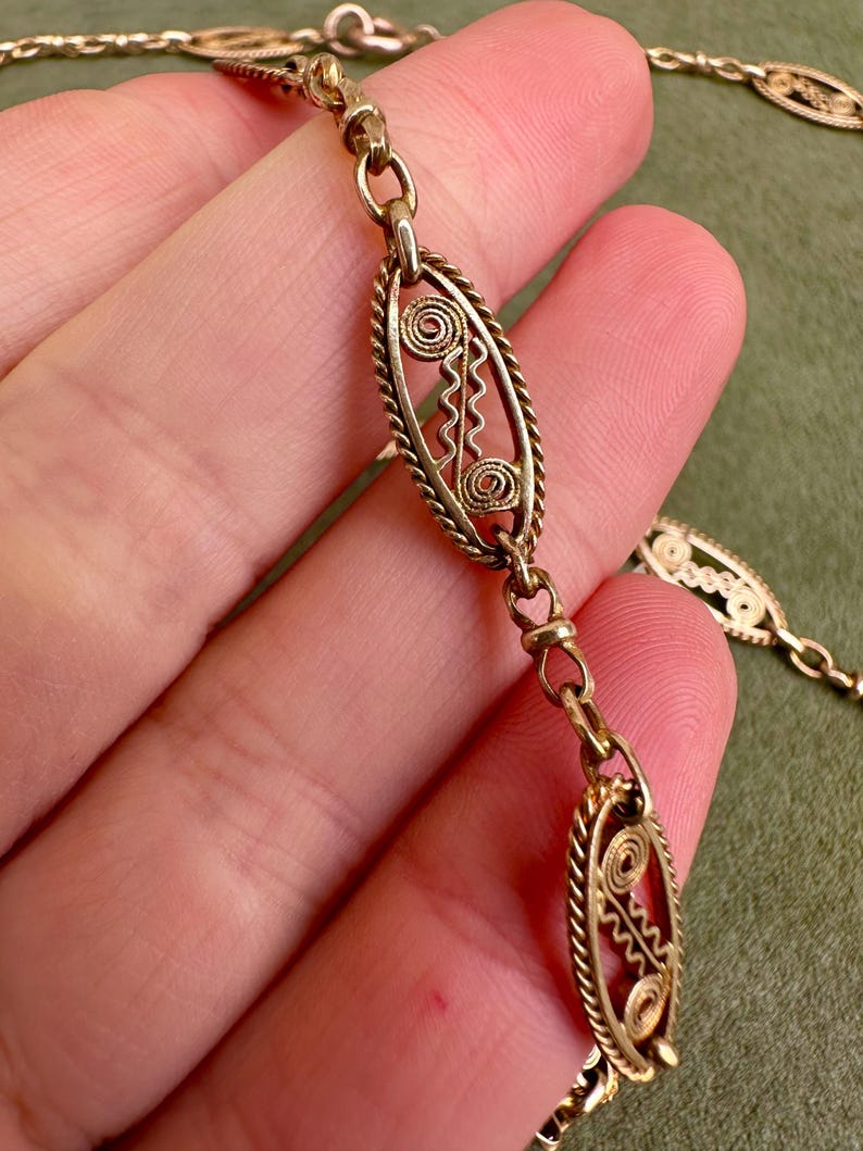 May include: A gold chain necklace with oval-shaped links featuring intricate filigree designs. The links are connected by smaller, round links.