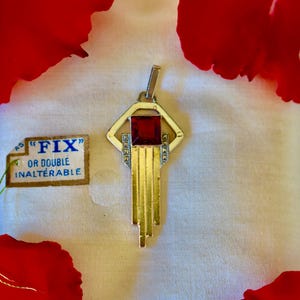 May include: Art Deco pendant featuring a red rectangular stone in a silver-tone metal frame. The pendant has gold-tone vertical bars and decorative accents. A small tag with the word "FIX" is visible.