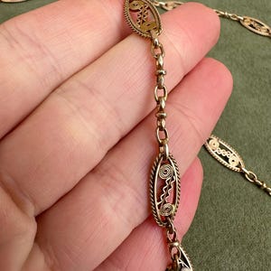 May include: A gold chain necklace with oval-shaped links featuring intricate filigree designs. The chain is delicate and has a vintage aesthetic.