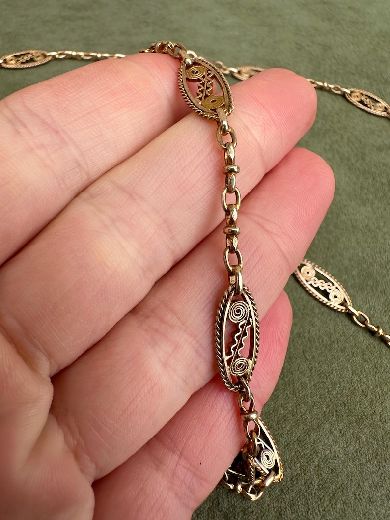 May include: A gold chain necklace with oval-shaped links featuring intricate filigree designs. The chain is delicate and has a vintage aesthetic.