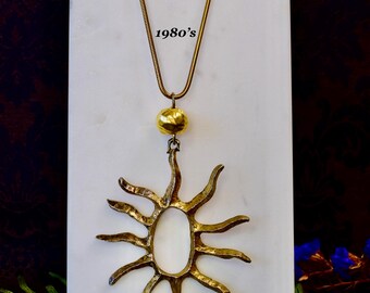 Puede incluir: Un collar de oro con un colgante en forma de sol. El colgante está hecho de metal texturizado y tiene una gran abertura ovalada en el centro. El collar es del diseñador de joyas francés Edouard Rambaud y data de la década de 1980.