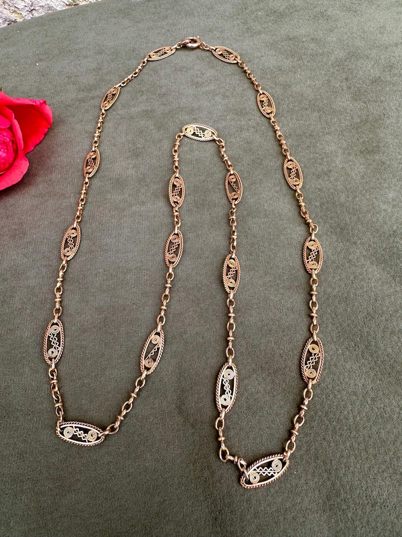 May include: A gold chain necklace with oval-shaped links. Each link is intricately designed with a filigree pattern.